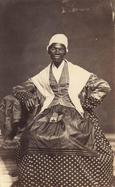 <p>Black and white photograph of a middle aged Black woman wearing glasses, a white cap and shawl, a polka-dotted dress, and a jacket and apron. She sits facing the camera with one hand on her hip and her other elbow resting on a small table. In her lap is a small framed photograph of her grandson.</p>